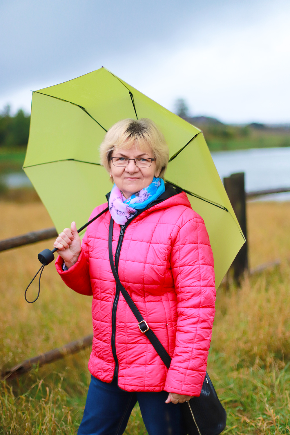 Natural Pharmaceuticals ImuBerin Középkorú nő napsárga esernyővel, rózsaszín kabátban, kék és lila sállal, vidéki őszi tájon séta közben.