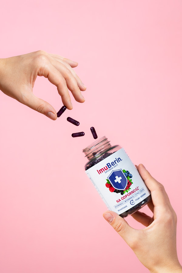 Natural Pharmaceuticals ImuBerin Kéz, amelyben nyitott csomagolású ImuBerin® immunerősítő étrend-kiegészítő látható, amelyből a másik kéz lila kapszulákat önt ki, rózsaszín háttér előtt.